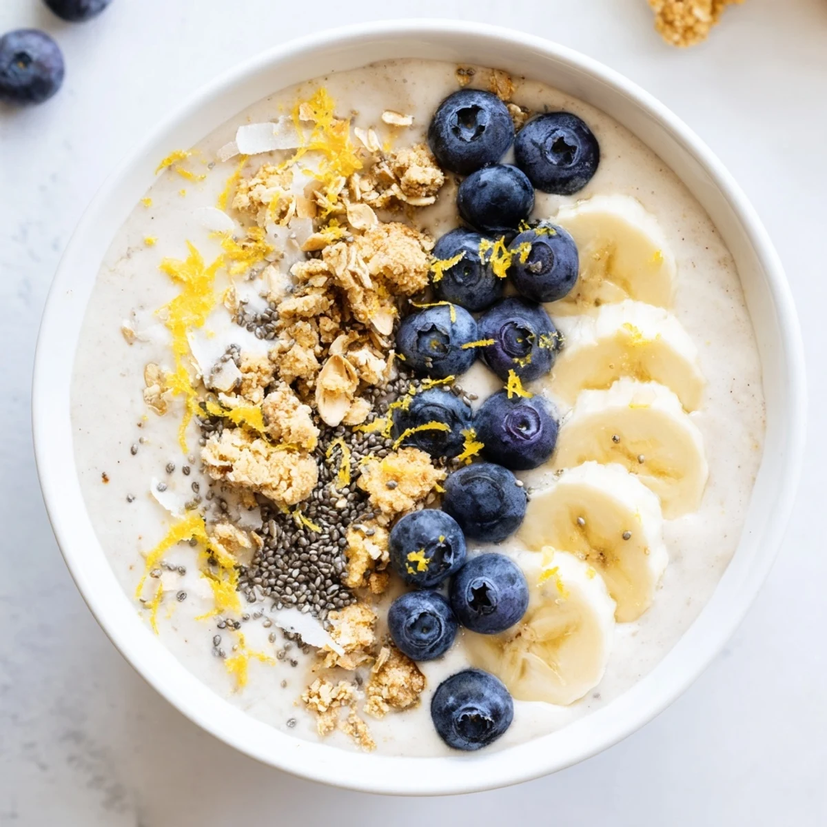 Close-up of a thick, purple Lemon Blueberry Smoothie Bowl in a white bowl, topped with fresh blueberries, banana slices, granola, chia seeds, and coconut flakes.