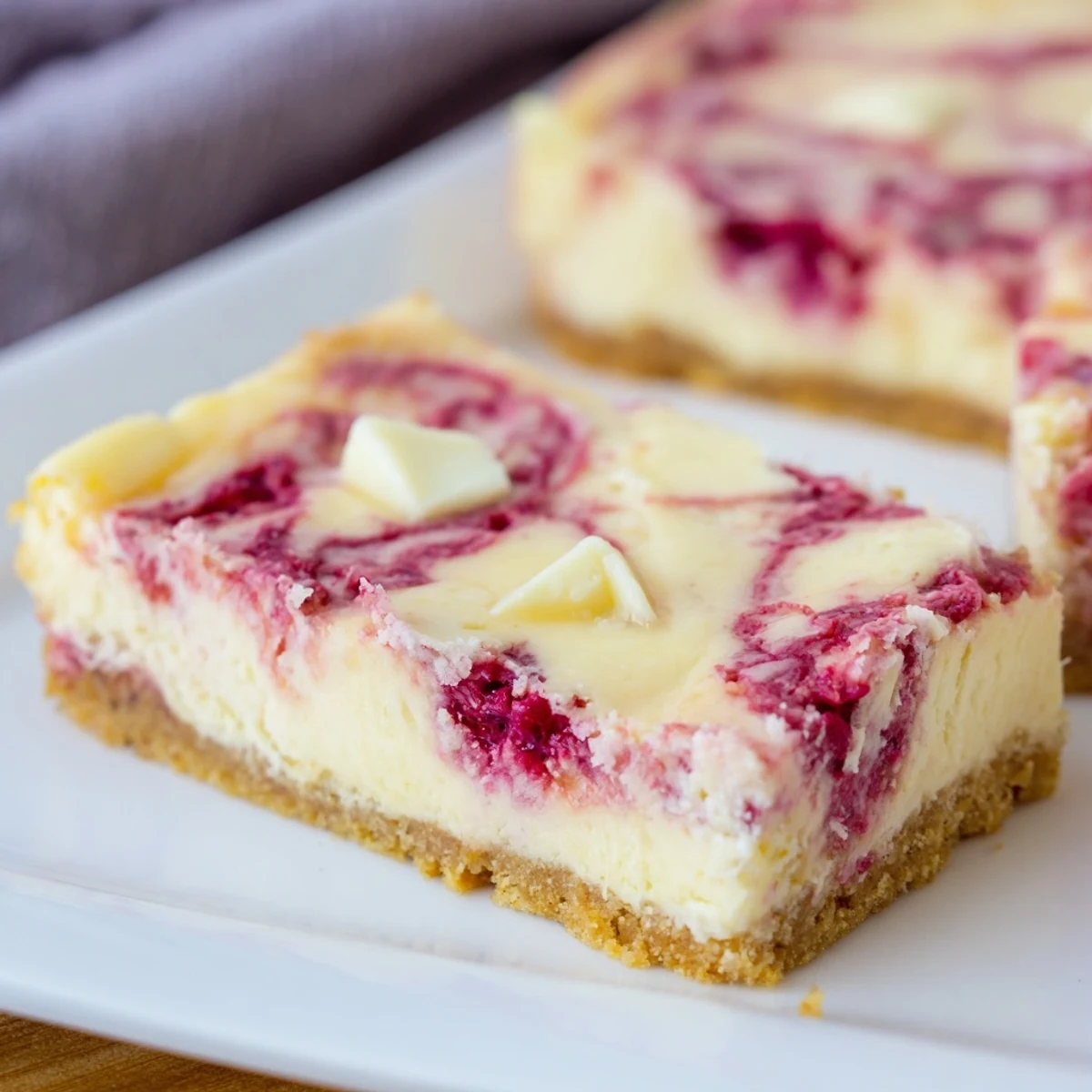 Slice of creamy raspberry white chocolate cheesecake bars with a buttery graham cracker crust and vibrant red swirls.