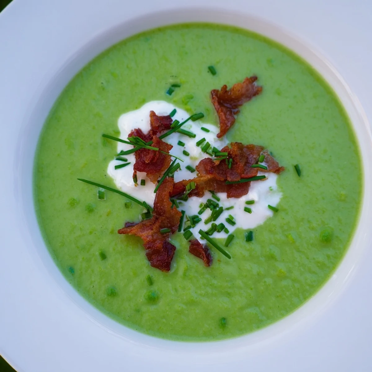 A close-up of vibrant Spring Pea Soup in a white bowl, topped with crispy turkey bacon and fresh chives.