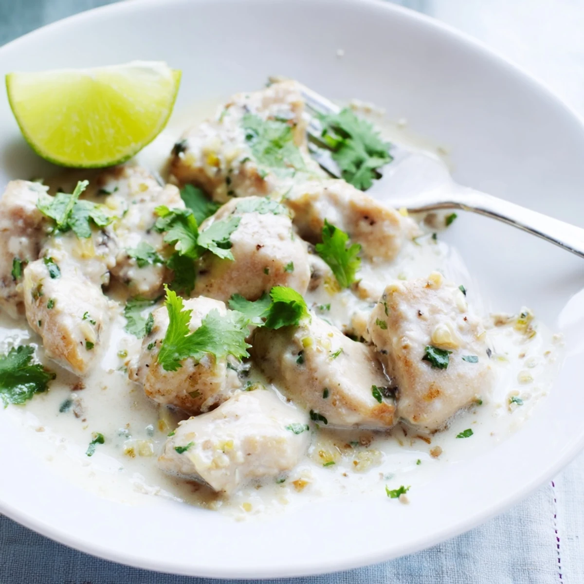 Golden coconut lime chicken swimming in creamy white sauce with vibrant green cilantro garnish and fresh lime wedges on a white plate.